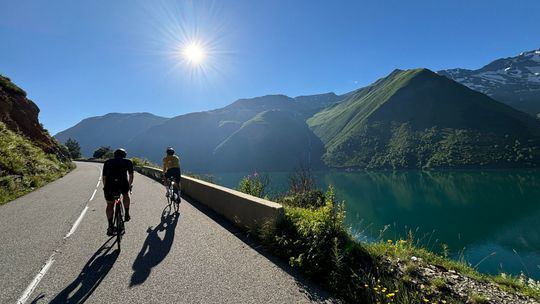 Two cyclists riding along a sunlit mountain road next to a turquoise lake
